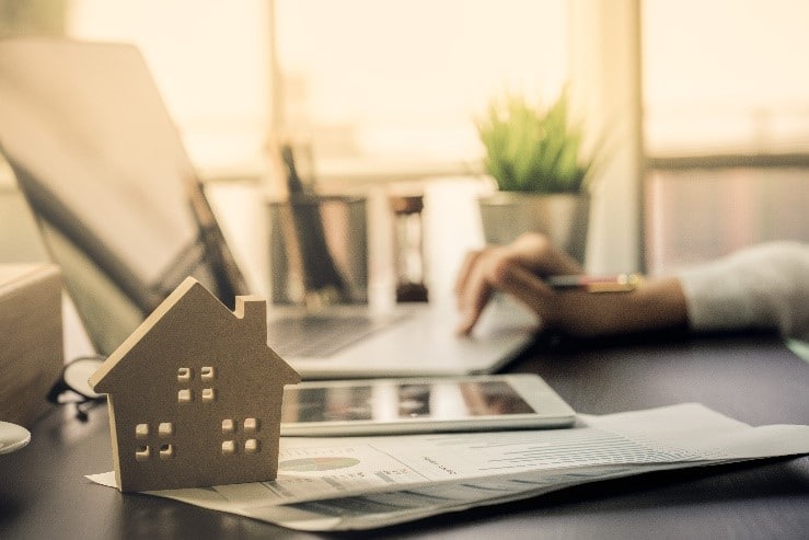 A photo of a person on their computer with a replica of a small house in the foreground. There is a plant in the background that is out of focus. 