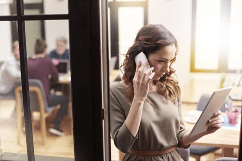 A Businesswoman Makes a Phone Call While Looking at Her Tablet While Other Co-Workers are at a Table in the Background