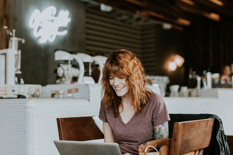 A Smiling Woman Sits in a Coffee Shop While Reading Something on the Screen of Her Laptop